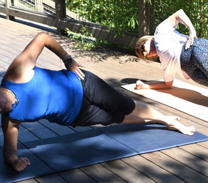 Two women doing side-plank yoga core workout on mats on a sunny wooden deck patio with lush green foliage