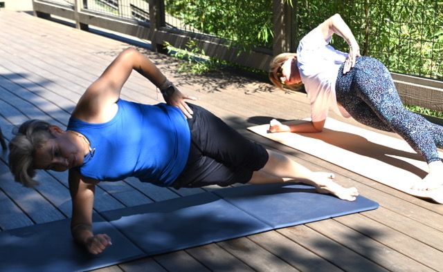 Two women doing side-plank yoga core workout on mats on a sunny wooden deck patio with lush green foliage