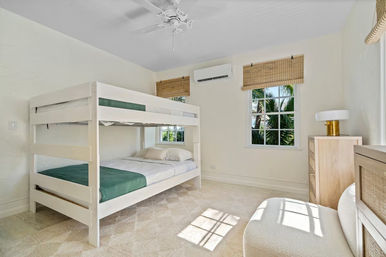 Airy coastal bedroom with white bunk bed, green bedding, bamboo window shades framing a palm-tree view, light wood dresser and ceiling fan