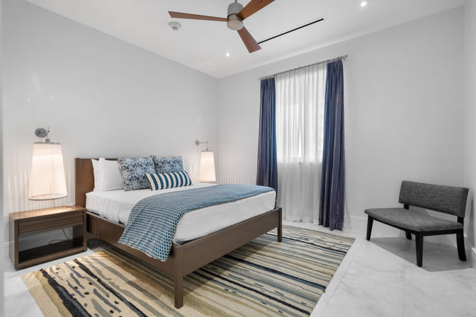 Bright modern bedroom with blue accents: wooden platform bed topped with patterned pillows and a blue throw, navy curtains over a sheer window, striped area rug, ceiling fan, and pendant bedside lamps.