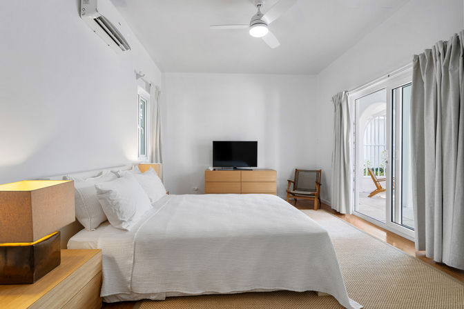 Sunlit modern minimalist bedroom with king bed in white linens, wooden nightstand and dresser, ceiling fan and wall air conditioner, and sliding glass doors opening to a small patio with outdoor chairs.