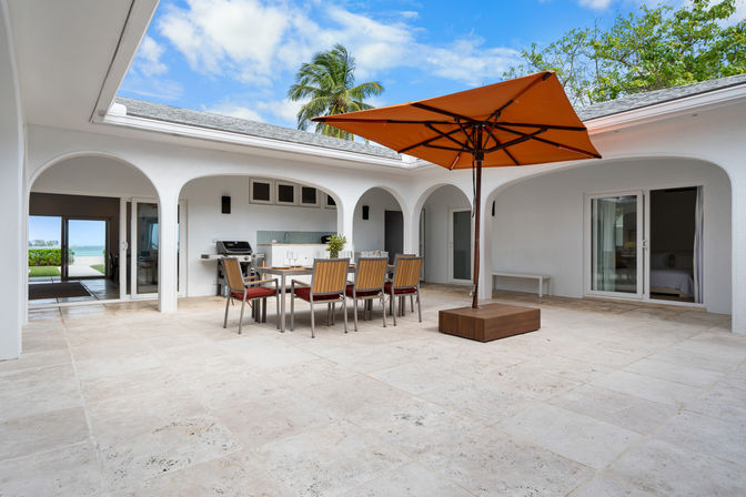 Sunny tropical courtyard patio with white stucco arches, light stone tile floor, wooden dining table and chairs beneath a large orange umbrella, built-in outdoor grill and kitchenette, sliding glass doors to rooms and a palm tree with ocean view beyond.
