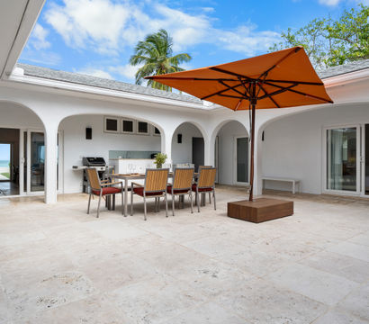 Sunny tropical courtyard patio with white stucco arches, light stone tile floor, wooden dining table and chairs beneath a large orange umbrella, built-in outdoor grill and kitchenette, sliding glass doors to rooms and a palm tree with ocean view beyond.