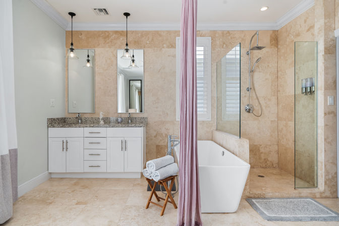 Spa-like luxury bathroom with beige marble tile, white double vanity with granite countertop and pendant lights, freestanding soaking tub with folded towels on a wooden stool, and glass-enclosed walk-in shower with shuttered window.