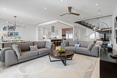 Bright modern open-concept living room and kitchen with matching gray sofas, round dark-wood coffee table topped with green apples, light area rug, stainless-steel appliances, island with bar stools, and a floating staircase with glass railing.