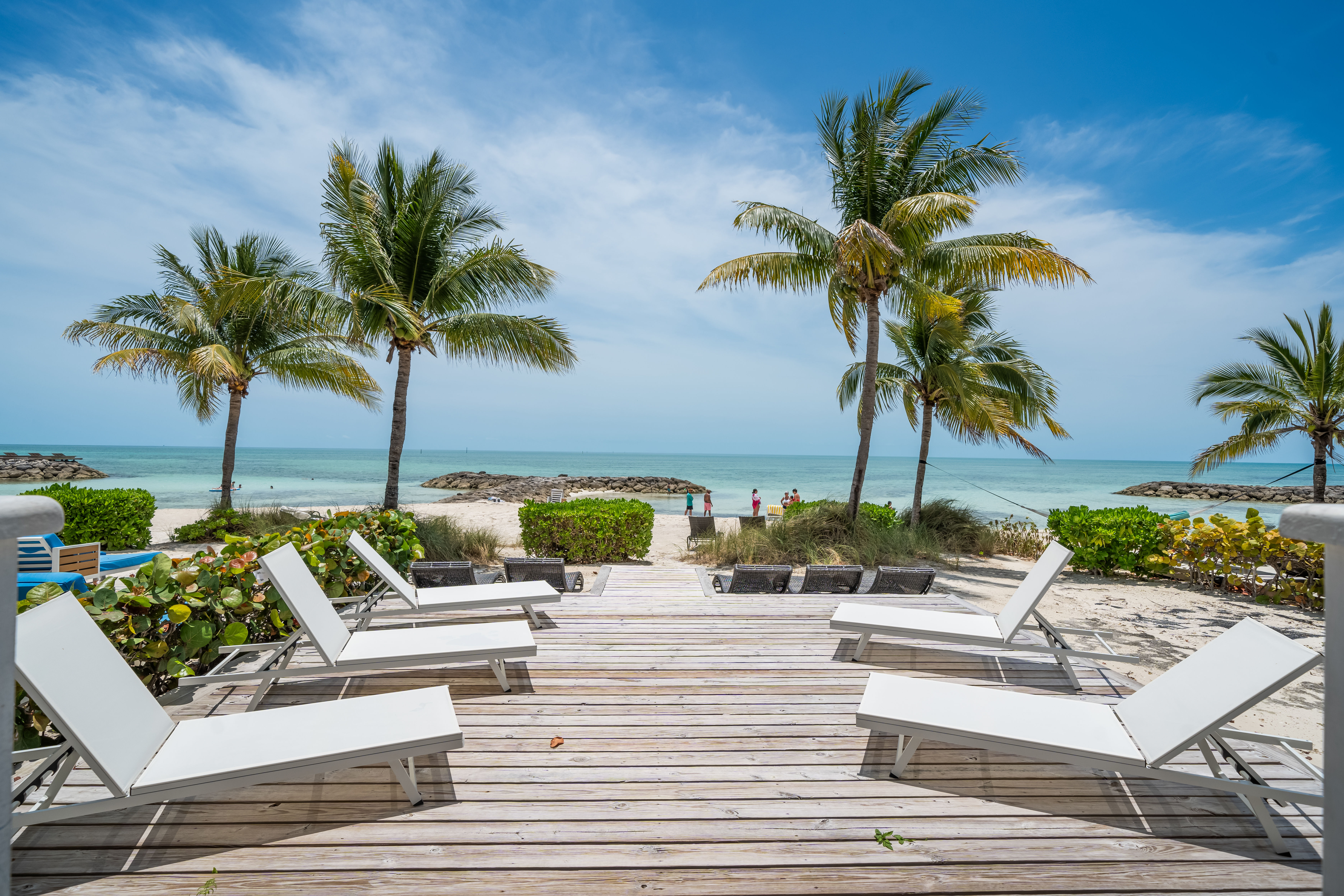 Sunny tropical beach with wooden deck and white lounge chairs, palm trees framing turquoise ocean and white sand under a blue sky.