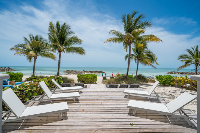 Sunny tropical beach with wooden deck and white lounge chairs, palm trees framing turquoise ocean and white sand under a blue sky.