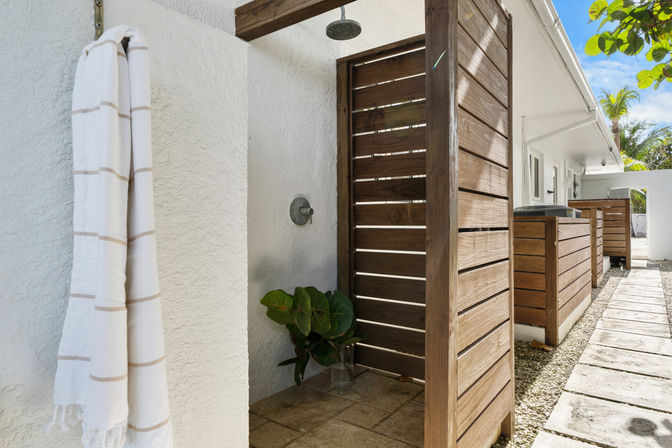 Outdoor beachside shower with wooden slatted privacy panels, white stucco wall, striped towel, tropical plant and stone tile pathway
