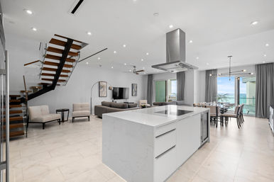 Modern open-concept beachfront condo interior with a large white marble kitchen island and cooktop, floating wood-and-glass staircase, gray living room seating, dining table by floor-to-ceiling windows opening to an ocean-view balcony, neutral tile floors and recessed lighting.