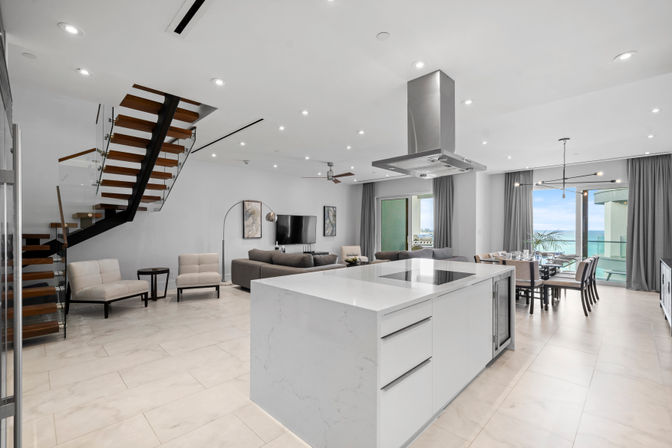 Modern open-concept beachfront condo interior with a large white marble kitchen island and cooktop, floating wood-and-glass staircase, gray living room seating, dining table by floor-to-ceiling windows opening to an ocean-view balcony, neutral tile floors and recessed lighting.