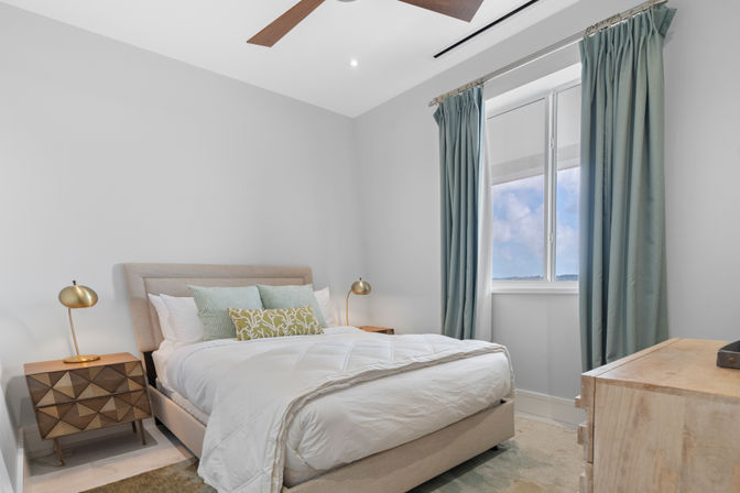 Cozy modern bedroom interior with upholstered bed and white bedding, patterned throw pillows, teal floor-length curtains framing a bright window view, geometric wood nightstand with brass lamps, and ceiling fan.