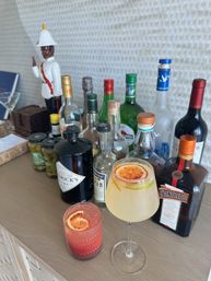 Home bar scene on a wooden sideboard with assorted spirits bottles and jars of olives, two cocktails β a pink fizzy drink and a pale citrus spritz β each garnished with a blood orange slice, and a white-uniform decorative figurine in the background.
