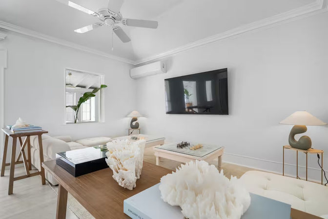 Bright coastal-style beach house living room with white walls and ceiling fan, wall-mounted TV, tufted white sectional, glass-top coffee table with chess set, decorative coral sculptures on a wooden console, and a tropical potted plant by the window — airy modern coastal vibe.
