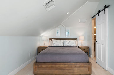 Cozy attic-style bedroom with a sloped ceiling, wooden platform bed dressed in gray bedding, matching nightstands with lamps, two framed prints above the headboard, and a white sliding barn door to a bathroom.