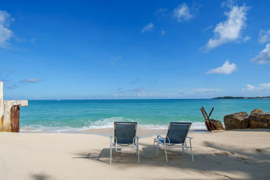 Two empty lounge chairs on a white sandy tropical beach facing turquoise ocean and clear blue sky, with a weathered pier and rusted metal near a rocky shoreline