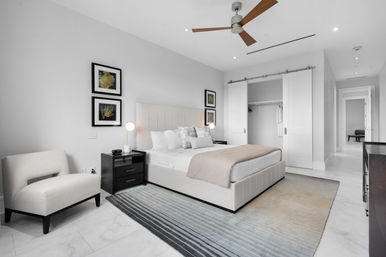 Bright modern master bedroom interior with an upholstered platform bed, plush pillows and blush throw, black nightstands with lamps, cream accent chair, striped area rug, ceiling fan, and sliding closet doors.