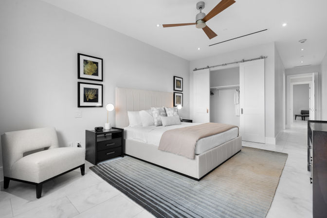 Bright modern master bedroom interior with an upholstered platform bed, plush pillows and blush throw, black nightstands with lamps, cream accent chair, striped area rug, ceiling fan, and sliding closet doors.