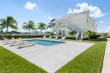 Sunny waterfront getaway: white two-story beach house with a rectangular pool, row of sun loungers, palm trees and a marina with boats under a clear blue sky.