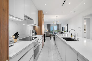 Bright open-concept modern kitchen with long white marble island and dual chrome sinks, integrated stainless-steel appliances, wood-accent cabinets, knife block and toaster, flowing into a dining and living area with large windows and balcony view.