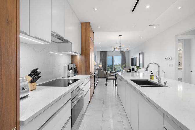 Bright open-concept modern kitchen with long white marble island and dual chrome sinks, integrated stainless-steel appliances, wood-accent cabinets, knife block and toaster, flowing into a dining and living area with large windows and balcony view.