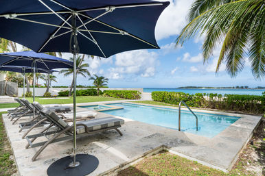 Tropical beachfront pool with striped chaise lounges under navy umbrellas, palm trees framing a turquoise ocean and blue sky.