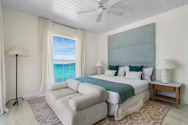 Sunny coastal ocean-view bedroom with king bed, teal upholstered headboard and matching runner and pillows, plush chaise at the foot, large white-curtained window overlooking turquoise sea, ceiling fan, rattan nightstand with lamp, and geometric rug on light wood floors.