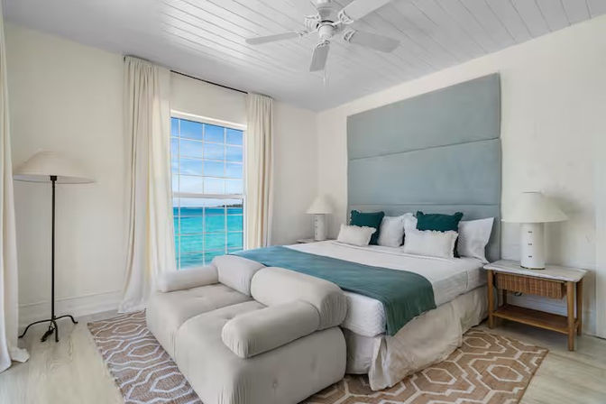 Sunny coastal ocean-view bedroom with king bed, teal upholstered headboard and matching runner and pillows, plush chaise at the foot, large white-curtained window overlooking turquoise sea, ceiling fan, rattan nightstand with lamp, and geometric rug on light wood floors.