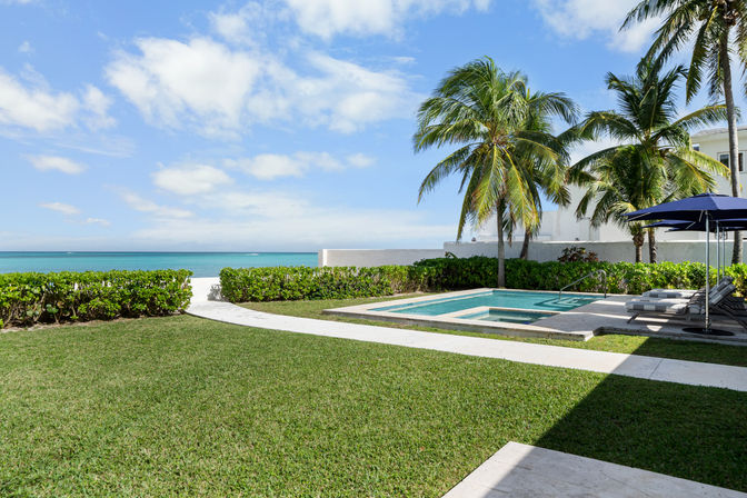 Oceanfront backyard with palm trees, private pool and spa, sun loungers under a navy umbrella, manicured lawn and a paved path leading to turquoise beach and blue sky