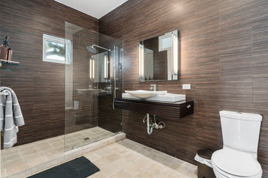 Spa-like modern bathroom interior with frameless glass walk-in shower and rain showerhead, floating vanity with vessel sink and illuminated mirror, wood-look tile walls and beige stone floor
