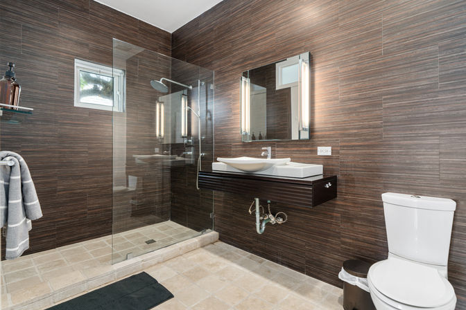 Spa-like modern bathroom interior with frameless glass walk-in shower and rain showerhead, floating vanity with vessel sink and illuminated mirror, wood-look tile walls and beige stone floor