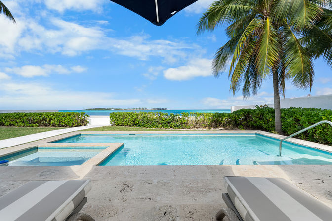 Tropical beachfront pool with turquoise water, attached spa, striped lounge chairs, palm tree and umbrella framing a clear blue ocean and sky