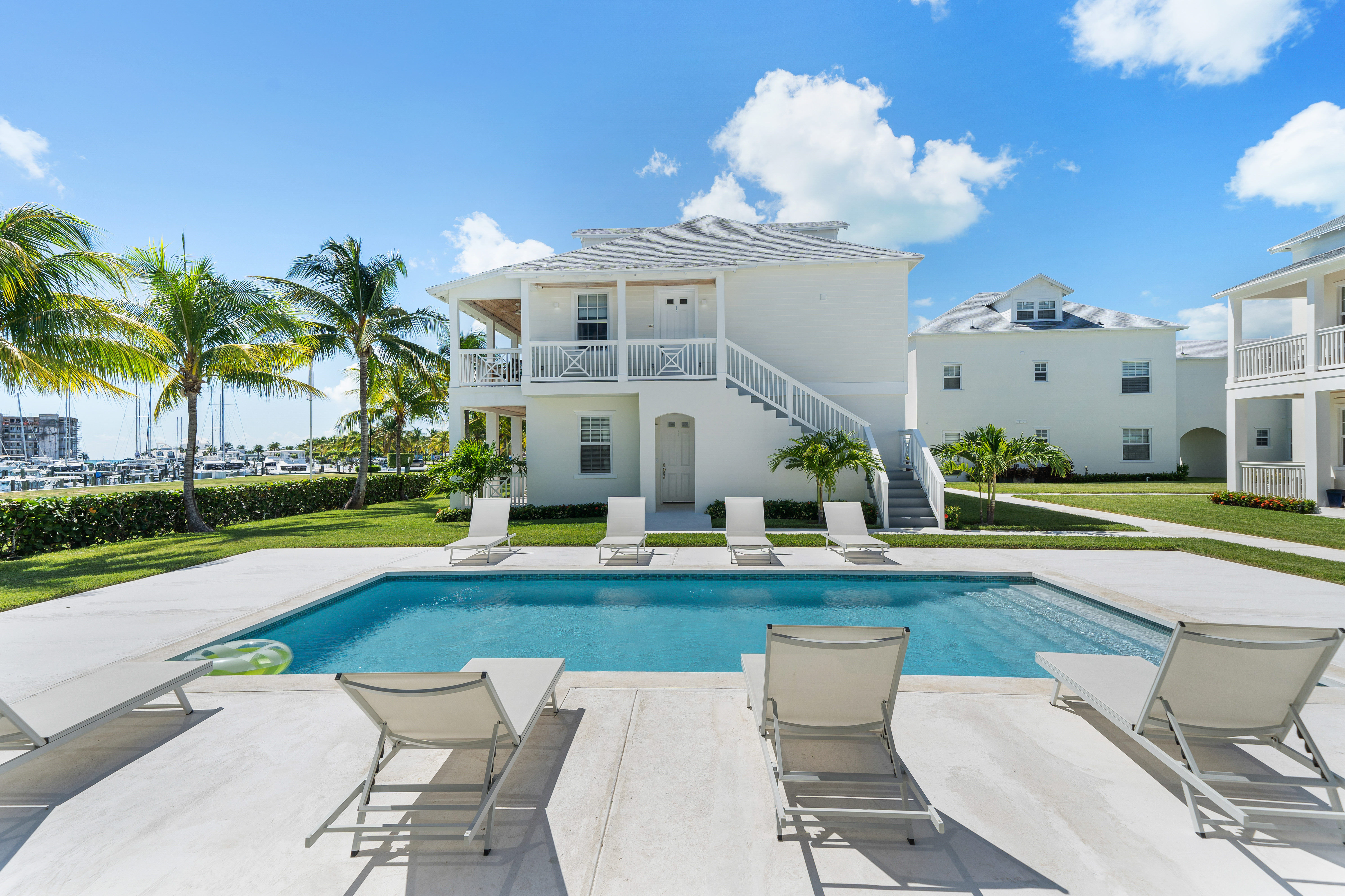 Tropical waterfront vacation property with bright white coastal duplex, sparkling pool, sun loungers, palm trees and marina in the background