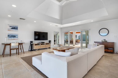 Bright modern living room with large white sectional, vaulted ceiling and ceiling fan, wall-mounted TV above a rattan media console, small desk with ocean prints, sliding glass doors opening to a sunlit patio with orange umbrellas, and beige tile floors.