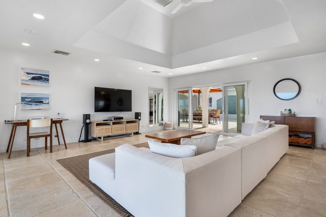 Bright modern living room with large white sectional, vaulted ceiling and ceiling fan, wall-mounted TV above a rattan media console, small desk with ocean prints, sliding glass doors opening to a sunlit patio with orange umbrellas, and beige tile floors.