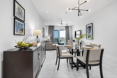 Modern open-plan living and dining room in a bright condo — glass dining table with gray upholstered chairs, dark sideboard with fruit bowl, neutral sofa, balcony view and contemporary chandelier