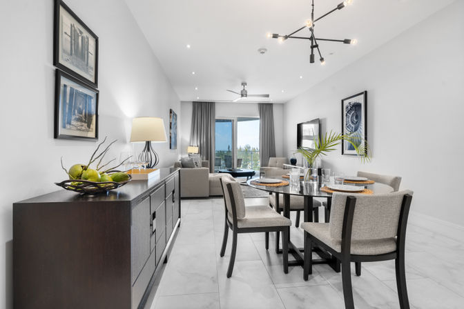 Modern open-plan living and dining room in a bright condo — glass dining table with gray upholstered chairs, dark sideboard with fruit bowl, neutral sofa, balcony view and contemporary chandelier