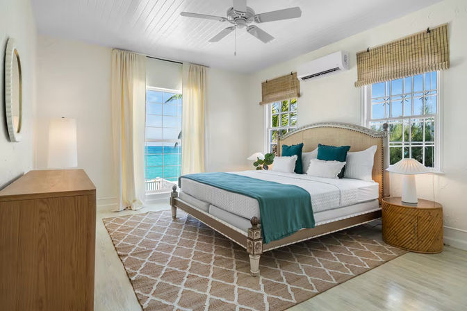 Bright beachfront bedroom with king bed, teal throw and pillows, rattan nightstand and woven shades, large windows framing turquoise ocean and palm tree.