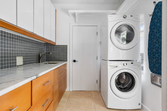 Bright compact modern laundry room with stacked white washer and dryer beside a marble countertop, light wood cabinets, dark gray square tile backsplash, sink, and wall-mounted blue ironing board.