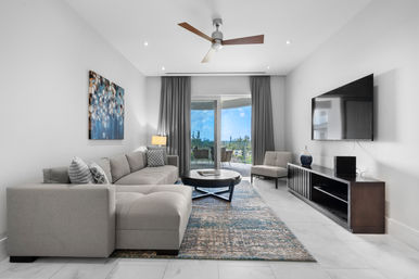 Bright modern living room with gray sectional sofa, patterned rug, round coffee table, wall-mounted TV, ceiling fan, and sliding glass doors leading to a balcony with blue-sky greenery view.