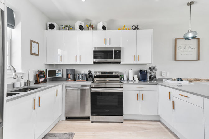 Bright modern white U-shaped kitchen with glossy cabinets and brass pulls, stainless steel oven, microwave and dishwasher, gray countertops, coffee station and toaster, pendant light and beach-themed wall art.