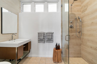Bright, spa-like modern bathroom with clerestory windows, glass-enclosed walk-in shower and wood-look tile, floating vanity with white sink, gray striped towels on the wall, and a rustic wood stool holding amber soap dispensers.
