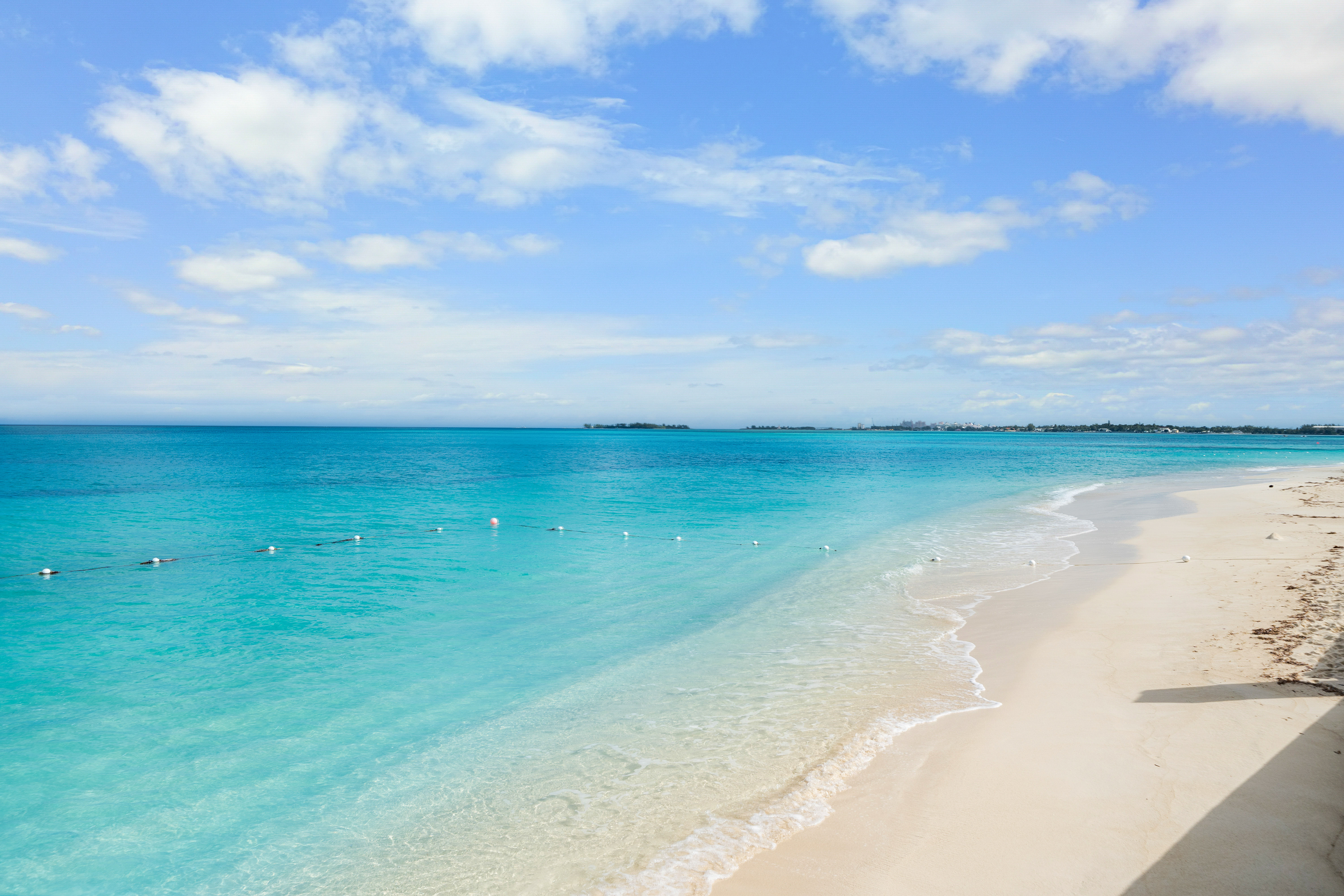 Tropical turquoise Caribbean beach with powder-white sand, gentle waves, a line of floating buoys, and a bright blue sky dotted with fluffy clouds