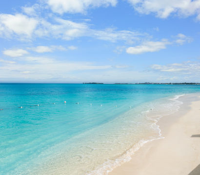 Tropical turquoise Caribbean beach with powder-white sand, gentle waves, a line of floating buoys, and a bright blue sky dotted with fluffy clouds