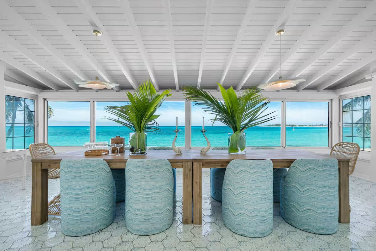 Sunny oceanfront dining room with a long wooden table, aqua wave-patterned stools, panoramic windows framing turquoise sea and palm trees, and tall palm fronds in glass vases as centerpieces.