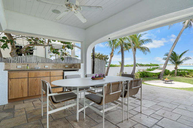 Covered beachfront patio with white arched ceiling and fan, outdoor dining table with wicker chairs, built‑in kitchenette and tiled floor opening to palm trees, hammock, fire pit and turquoise ocean view in a tropical seaside backyard.