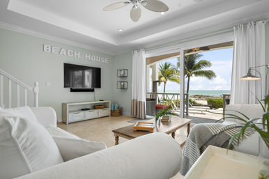 Bright beachfront living room with white sofas, wooden coffee table and wall-mounted TV, sliding glass doors opening to a palm-lined patio and ocean view