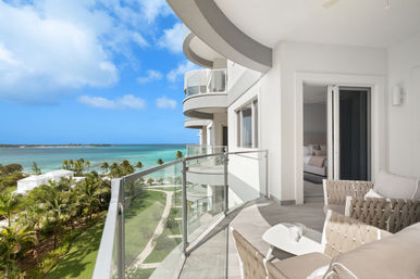 Sunny beachfront condo balcony with glass railing, cozy woven chairs and table overlooking palm-lined grounds and turquoise ocean under a blue sky