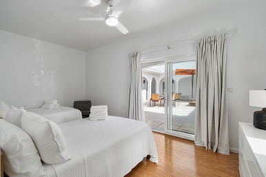 Sunlit minimalist bedroom with white linens, ceiling fan and hardwood floor, sliding glass doors opening to a courtyard patio with outdoor dining table and umbrella.