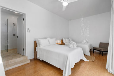 Bright minimalist guest room with two white-made beds, wooden nightstand and lamp, rolled towels, hardwood floors, ceiling fan, and en-suite tiled shower.