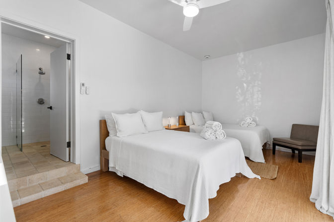 Bright minimalist guest room with two white-made beds, wooden nightstand and lamp, rolled towels, hardwood floors, ceiling fan, and en-suite tiled shower.